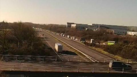 A traffic camera picture taken from above a motorway bridge. A long line of stationary traffic can be seen queuing up on the far side carriageway. 