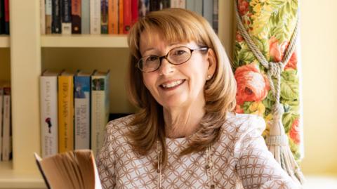 Erica James in a white and gold top sits in front of a book shelf with a novel in her hand