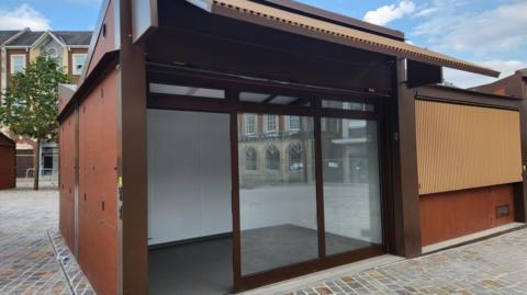 Brown rectangular building with full-length glass window and door on side, with market square visible to one side and in the window reflection.