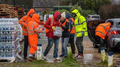 Seven people, six in hi-vis orange and one is hi-vis yellow are standing next to a pallet of bottled water