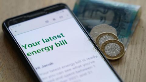 A mobile phone on a table, with an email open that says "your latest energy bill" in green writing.  On the table behind there is a five pound note and some coins.