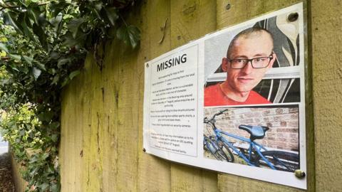 A missing poster for Martyn Richardson, which is pinned to a wooden fence. It has a picture of Mr Richardson, who is wearing black glasses and a red T-shirt. It also has a picture of his bike, which is blue. 