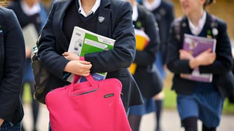 Secondary school-aged girls in uniforms are seen walking and holding school books to their chests. 