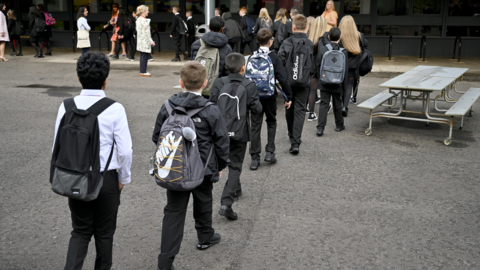 A line of school children carrying bags walking across a playground.
