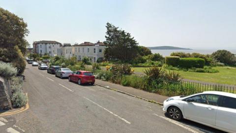 A Google Street View of Upper Kewstoke Road. It overlooks the sea, and has a large park area on the right hand side. Cars are parked along the road and in the distance are large Victorian seaside houses. 