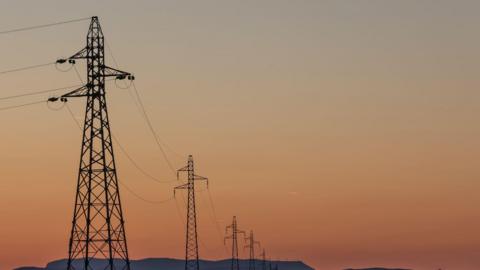 A line of tall metal electricity pylons stretching into the distance. These pylons are connected by long, thin power lines that arc slightly between each structure. The stock image has been taken at sunrise or sunset.