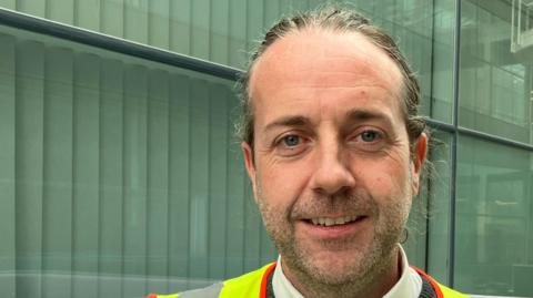 A head and shoulders picture of Colin Walsh wearing a high-vis jacket inside a building at Stansted Airport. His hair is swept back and he has facial stubble. He is wearing a white shirt under a grey crew-neck jumper.