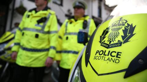 The front of a police vehicle with POLICE SCOTLAND written on it, and two officers standing in the background 