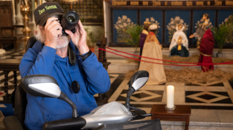 A man in a wheelchair, his face covered by a large camera lens, with a scene of the nativity in the cathedral behind him