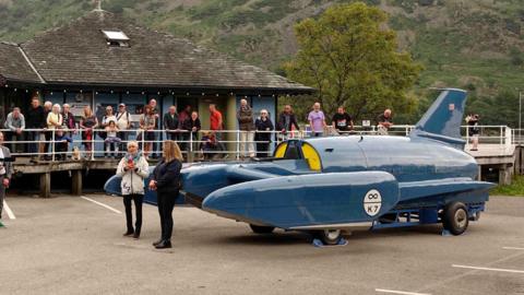 Gina Campbell and Rachel Bell, head of marketing and business development at Ullswater Steamers and Cumbria Tourism’s vice-chair, stand next to Bluebird - a jet-powered hydroplane - in the car park at Glenridding Pier. There are dozens of onlookers and fells can be seen towering above in the background.