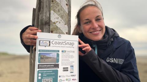 A lady with long brown hair tied back in a ponytail wearing a navy coat with GSNI written on it. She is standing beside a 'CoastSnap' poster which is pinned to a wooden post.