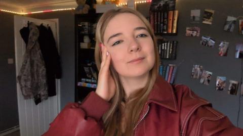Selfie image of Faith Best, who has long fair hair and is wearing a red jacket indoors. There are clothes hanging on the back of a white internal door. There are books on shelves and a series of photos placed onto a grey wall. 
