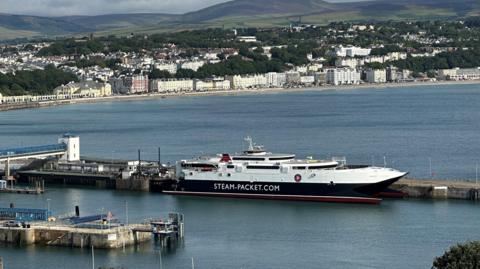 The Manannan ferry moored in Douglas Harbour with the beach and the promenade in the background and hills in the distance.