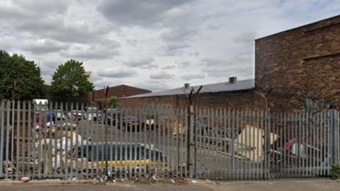 Cars are parked behind railings that are in the foreground. Trees are to the left of the image and a building goes from the middle to the right of the photo - these are also behind the railings.