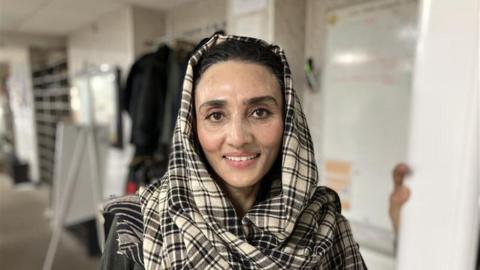 A woman in a black and white checked headscarf smiles at the camera. The background behind is blurred but she is standing in a white room with various objects in it.
