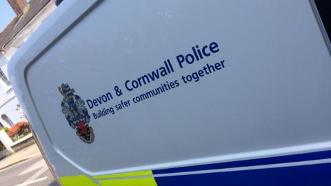 The side of a police van. It is blue and green in colour with a white gap - where the Devon and Cornwall Police logo is. Below the logo and text it says - 'Building safer communities together'