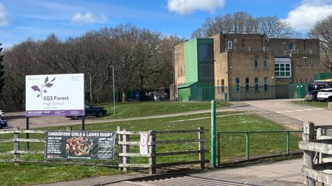 An aged secondary school building surrounded by woodland. There is a vehicle entrance with a cattle grid on the drive leading up to the school. There is a sign with the school name near the vehicle entrance and a rugby banner attached to the wooden fence. Cars can be seen in the car park surrounding the school.