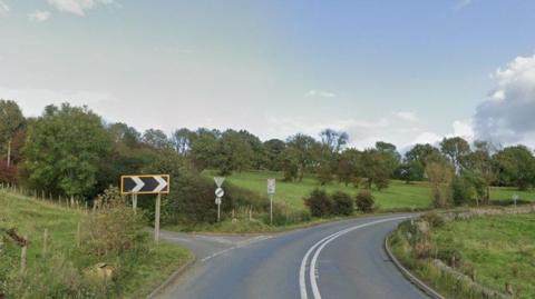 A section of the A523 in Waterhouses, near its junction with Stony Lane - on the left-hand side. The road is seen bending to the right, and there is chevron signage indicating the sharpness of the bend. It is a rural road, with fields and trees surrounding it, bounded by dry stone walls.