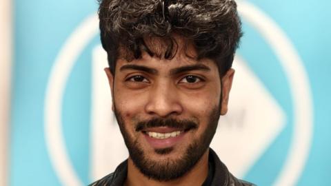 Man with moustache and beard smiles to camera with Reform UK branding behind him