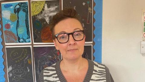 Emma Quinn is looking at the camera and standing in front of a wall with children's artwork hanging on it. She has brown hair, tied up and is wearing a dark green and cream striped jumper and black glasses. 