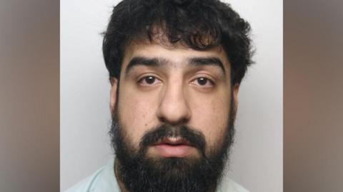 Custody photograph showing a person with dark hair and a full beard facing the camera against a plain light-coloured background.