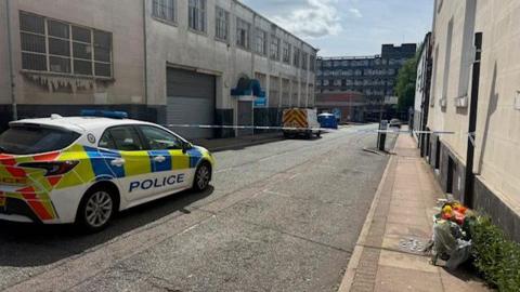 A police car parked in a narrow industrial street. Floral tributes can be seen at the opposite side of the road. Blue and white police tape cordons off the road ahead of the vehicle. Further up the roads another police vehicle can be seen and a blue police tent. 