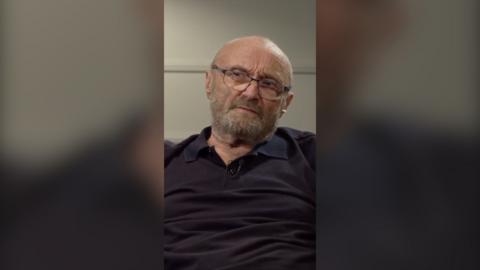 Phil Collins wear glasses and has a grey full beard and moustache. He is wearing a black, V-neck shirt.