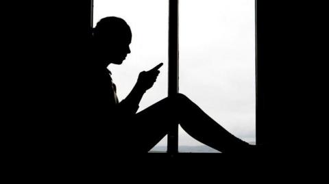 A high-contrast silhouette of a person sitting indoors in front of a large window. The person is positioned in profile view, facing toward the left side of the image. The outline of the figure is entirely dark, with no facial details visible due to the strong backlighting from the window behind.