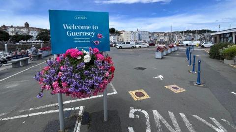 A sign says Welcome to Guernsey. It is surrounded by colourful flowers and sits at the edge of a car park on a pier.