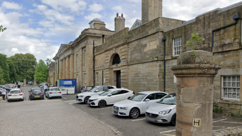 The exterior of Durham Crown Court. The building is made up of large sandstone bricks, and has a Georgian design with Roman columns at its entrance. There are several cars parked outside on a cobbled area of road. 