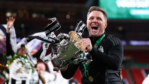 Eddie Howe on the pitch at Wembley, smiling holding the Carabao Cup trophy which has black and white ribbons hanging off it.