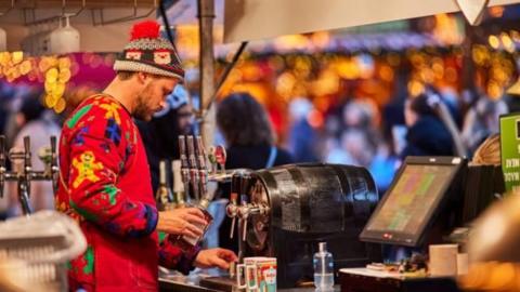 A bartender in a colourful Christmas jumper and wooly beanie stands behind a bar making drinks. 