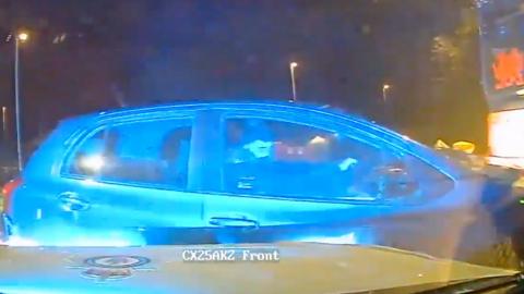 Grainy image from a police dash cam of a small black car, side on, illuminated in blue with the driver looking towards the police car. A police car bonnet can be seen in the foreground