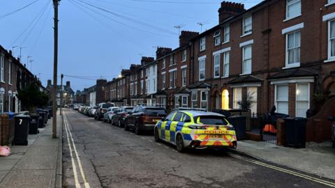 A lone police car on a residential road early in the morning. Homes line the road on either side. Other cars are parked on the road.