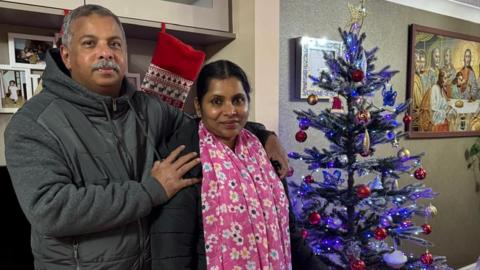 Jisha, a woman, wearing a black jacket and pink floral scarf around her neck, standing with her husband next to a purple Christmas tree.
