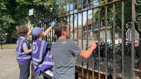 Three men working on railings outside York Minster. Two of them are wearing purple high vis jackets and one is wearing a grey t-shirt with a yellow logo on.