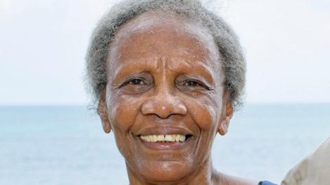 A woman with grey hair looking at the camera smiling. The sky and sea are in the background.