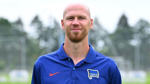 A bearded, shaven headed Gabor Ruhr faces the camera wearing a blue Hertha Berlin polo shirt