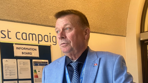 A man in a blue suit and tie looking to the side in a hallway with a notice board on the wall behind him.