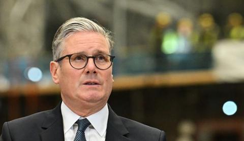 Keir Starmer stands in a dark suit, white shirt and blue tie; he wears round glasses and has short grey hair