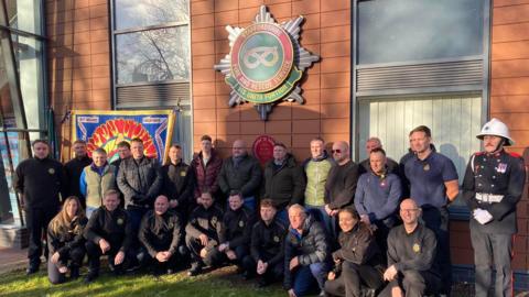 Friends and colleagues of Alec Elwell pose in front of red plaque