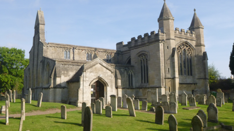 The outside of the church. The sky is blue with no clouds, the grass around the church is green and there are several gravestones on it. The church is a light stone colour and has large glass windows