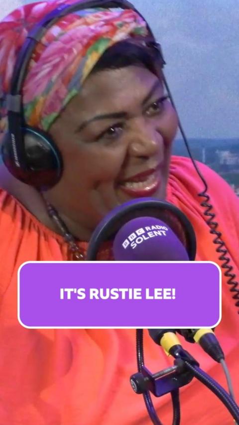 Rustie Lee wears a pink and green floral headscarf and bright orange blouse. She is sat in the radio studio with headphones over her head.