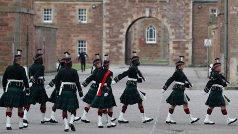 The soldiers are dressed in kilts and marching with guns resting on their shoulders. In the background are the walls of historic Fort George.