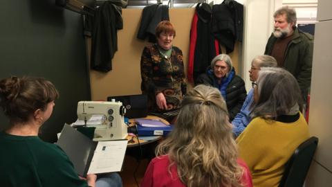 An image of six people sat at a table being taught how to use a sewing machine