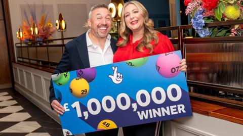 Richard Davies a Faye Stevenson-Davies yn gwenu ac wedi gwisgo'n smart. Mae'r ddau yn gafael mewn llun o siec mawr am £1 miliwn.