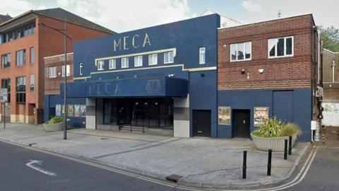 The venue pictured from across the road. The building is painted navy, in contrast to the brick next to it, and it has a large lobby entrance. It has the word MECA written on it twice in an Art Deco font. 