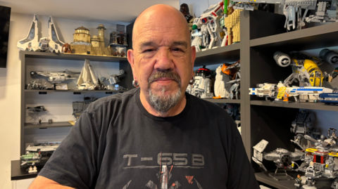 Pete Brierley is wearing a black T-shirt and standing in front of shelves with a range of model spacecraft.