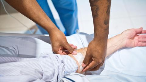 A healthcare worker securing medical tape over an IV tube on a patient's forearm. The patients is wearing a long-sleeved striped shirt and their arm rests on a hospital bed.