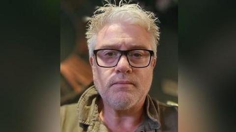 A man with white hair, dark glasses and a green jacket is looking at the camera.